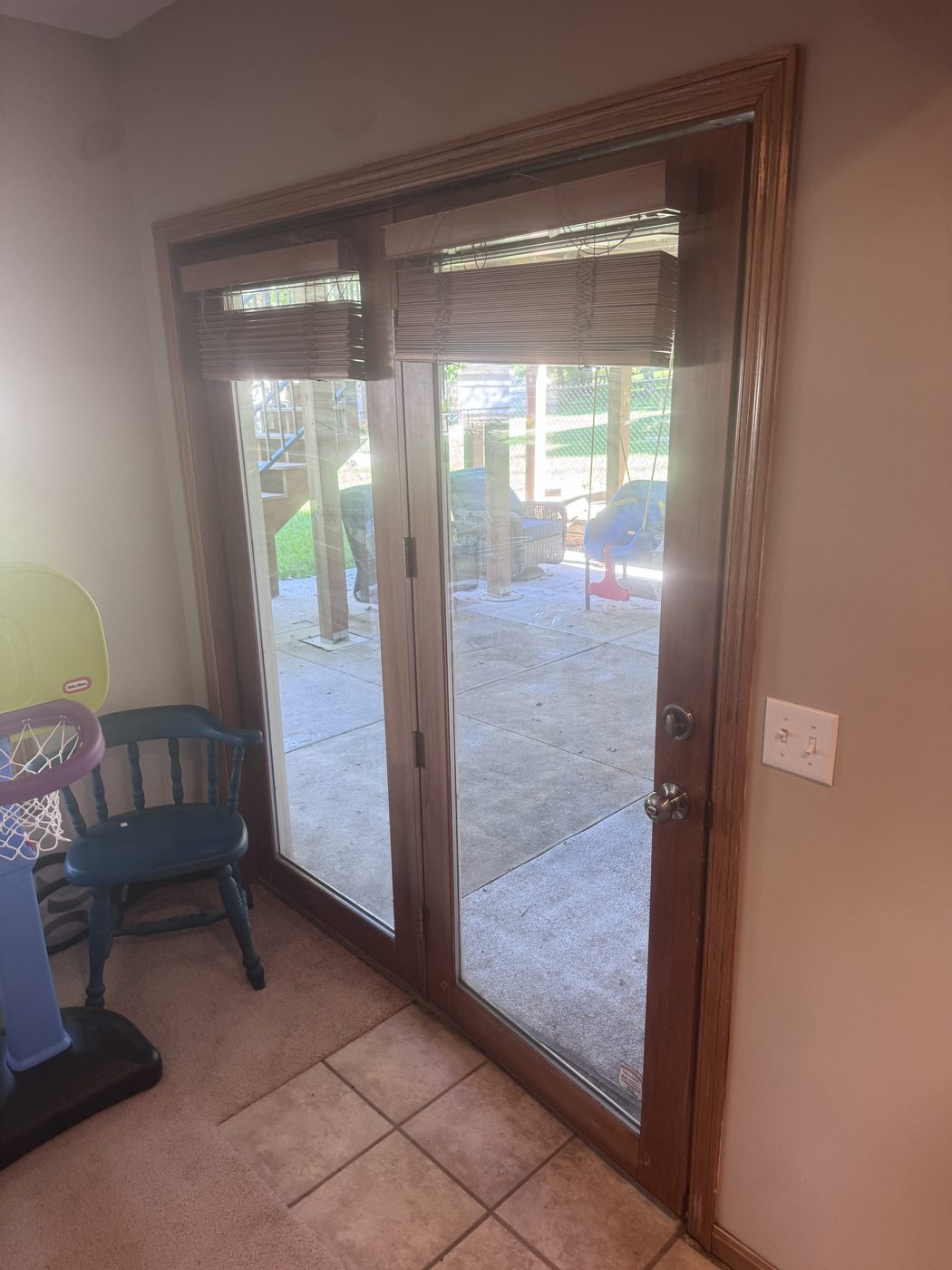 The original basement patio door from inside — a dated hinged unit with a fixed side panel, painted trim, and old mini blinds, opening onto the back patio